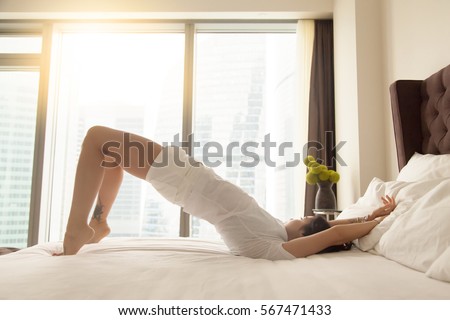 Young sporty attractive woman practicing yoga at home or hotel bedroom after waking up, stretching in dvi pada pithasana exercise, Glute Bridge, working out wearing white home wear, indoor full length