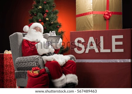 Santa Claus sitting on grey armchair with Sale sign and gift boxes, using tablet computer