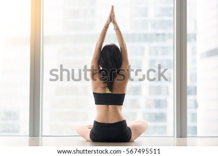 Rear view of woman practicing yoga, sitting in Padmasana exercise, Lotus pose, working out, wearing black sportswear, indoor full length, meditation session in the morning, floor window, back view 