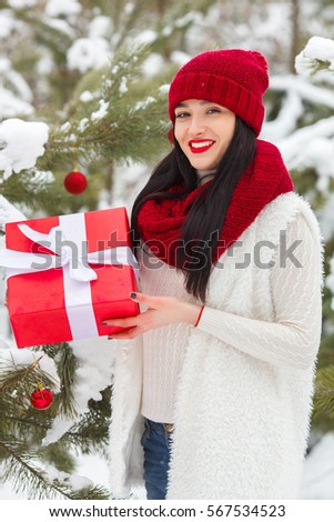 Young pretty girl smiling at camera, holding  presents or gifts in her hands. Beautiful woman looking forward, wearing red hat, scarf in the snow.