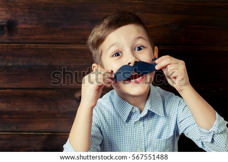 A kid with props for a photo booth. A child with the requisite mustache on wooden background. Event, holiday, party. Copy space.