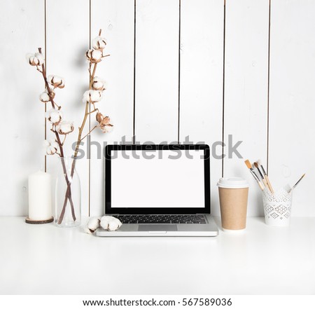 Stylish hipster workspace with open laptop computer, cup of coffee, painting brushes, candle and cotton plant branches in vase at home or studio.Desktop computer screen isolated. White desk mockup