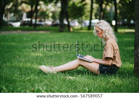 Beautiful young blonde woman sitting on the green grass in park near tree with her laptop in sunglasses. Student girl looking at the computer screen with her hands on keyboard.  