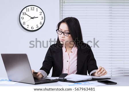 Pretty businesswoman working on the laptop computer with a clock on the wall in the office
