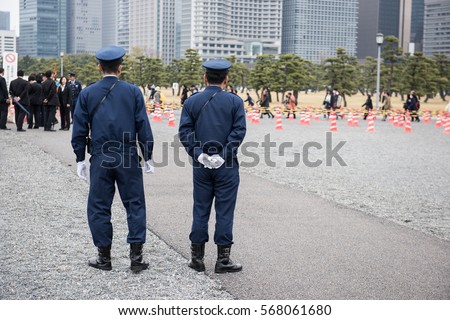 Here in the image two on duty Japanese police officers standing and watching people to maintain law and order in the area. And few people can be seen standing in a queue in the picture.
