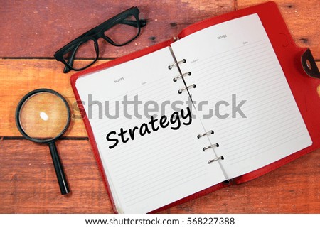 A concept image of an open red diary with spectacles and magnifying glass over a wooden background with a word Strategy