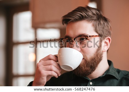 Picture of handsome bearded young man wearing glasses sitting in cafe while drinking coffee.