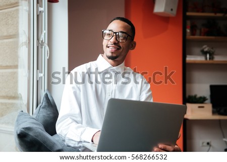 Picture of cheerful african businessman wearing glasses sitting near window and typing by laptop computer. Coworking.