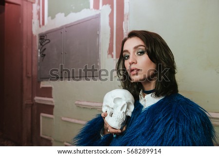 Picture of gorgeous young woman standing indoors at night. Holding artificial skull. Look at camera.