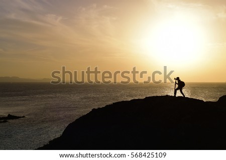 the silhouette of an unrecognizable young man taking a picture in front of the sea at dusk, against a colorful orange sky