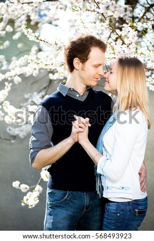 Tender couple in love under blooming cherry tree with white flowers in the park in spring. Happy girl and guy smile and hug outdoor, they are wearing in jeans, blue shirt, white jacket.
