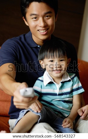 Father with young son on lap, holding TV remote control towards camera