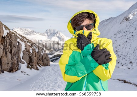 Woman feeling cold discomfort covering her mouth and face from the wind wearing gloves, hood, glasses and windstopper, at extreme snow elements environment on a snowy mountain with low temperature. Royalty-Free Stock Photo #568559758