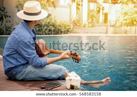 Romantic young man sitting on the pool at sunset with playing the guitar