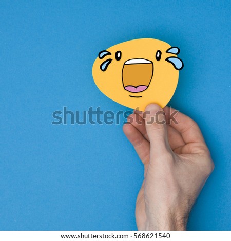 Laugh out loud emoji with tears. Male hand holding a yellow emotion face with a hand drawn expression