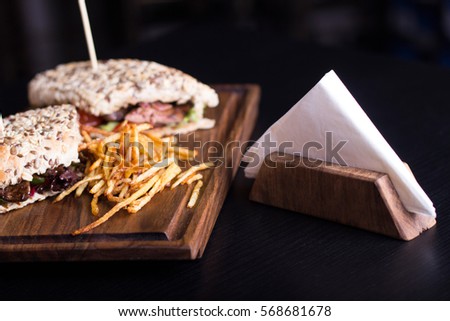 Tasty grilled sandwiches with a wooden stick stabbed trough them on a wooden plate. Meat sandwiches with lettuce, tomatoes and french fries on the side. Napkins on the side.