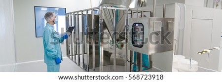 wide-screen picture, caucasian scientist in blue lab uniform make notes about big steel machine with control panel