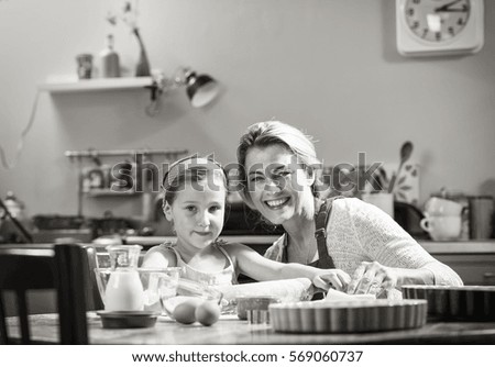 Looking at camera. A cheerful mother and her daughter having fun while making pastry in a kitchen. Black and white picture