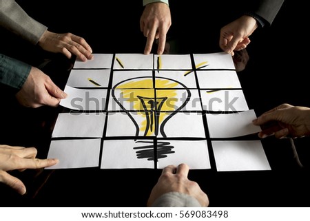 Brainstorming and teamwork concept with a team of seven businesspeople arranging white papers forming a yellow light bulb on a black table.