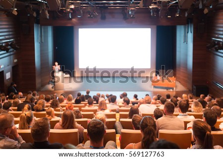 Audience at the conference hall. Speaker giving a talk in conference hall at business event. Business and Entrepreneurship concept. Lens focus on people in audience from rear. Royalty-Free Stock Photo #569127763