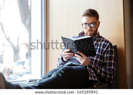 Young male student sitting on windowsill in library and reading book Royalty-Free Stock Photo #569289841