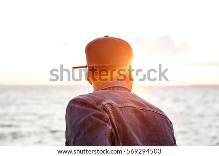 Back view photo of attractive african man wearing cap and dressed in jeans jacket looking at beach.