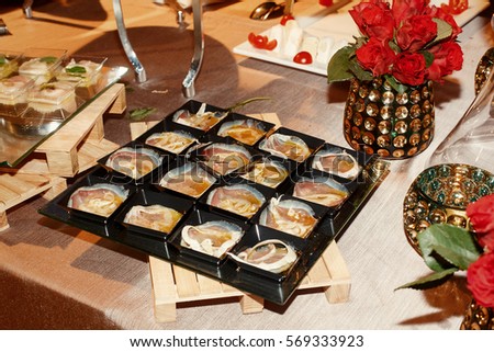 Little square black plates with snacks stand on dinner table among vases with roses