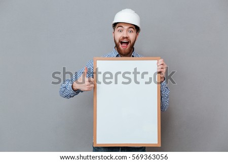 Picture of attractive young bearded builder holding copyspace board and make thumbs up gesture standing over grey background.