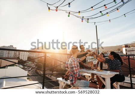 Outdoor shot of young people toasting drinks at a rooftop party. Young friends hanging out with drinks. Royalty-Free Stock Photo #569528032