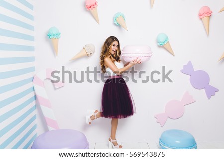 Joyful young woman in tulle skirt having fun with big macaroon on white background among sweets. Pastel colors, ice cream, happiness, cupcakes, smiling, surprised, playful