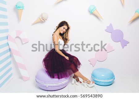 Sweet happy summer time of attractive fashionable young woman in tulle skirt sitting on huge macaroon on white background. Pastel colors, sweets, delicious, enjoying, happiness, smiling, relaxing