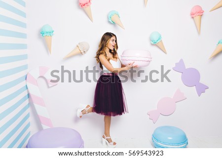 Funny surprised attractive young woman holding huge macaroon on white background among ice cream. Happy sweet moments, expressing positivity, diet concept, summer time