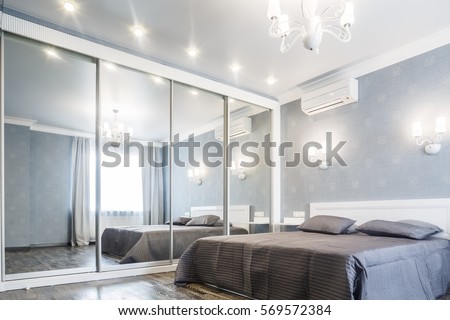 Contemporary bedroom design in a luxurious apartment. Hardwood floor, painted blue walls, white chandelier, rolling door wardrobe with mirrors. Royalty-Free Stock Photo #569572384