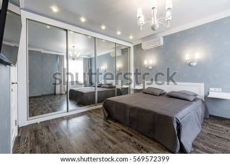 Contemporary bedroom design in a luxurious apartment. Hardwood floor, painted blue walls, white chandelier, rolling door wardrobe with mirrors. Royalty-Free Stock Photo #569572399