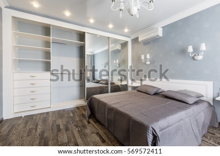 Contemporary bedroom design in a luxurious apartment. Hardwood floor, painted blue walls, white chandelier, rolling door wardrobe with mirrors. Royalty-Free Stock Photo #569572411