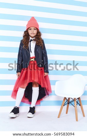 Beautiful cute pretty little girl baby blond curly hair fun to play in the children room blue stripe background chair wear cap skirt blouse jacket style fashion clothing collection daughter friend.
