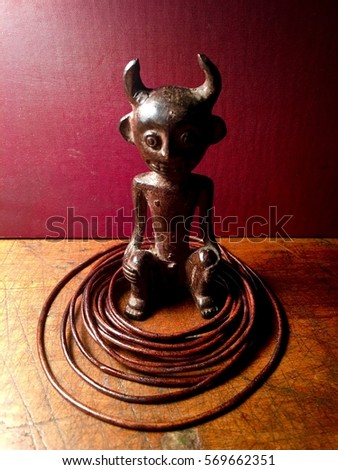 Statue of horned figure with loose leather bindings on a scarred wood surface against a dark red background. 
