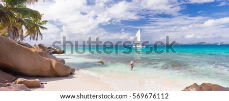 Woman wearing long floral summer dress and beach hat on Anse Patates beach on La Digue Island, Seychelles. Summer vacations on picture perfect tropical beach concept.