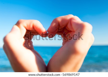 Composition finger frame- young woman's hands doing heart shape on seascape horizon. Close-up vibrant multicolored summertime horizontal outdoors inspirational image on blue sky background.