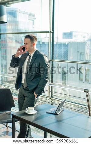 Young businessman in suit calling with smartphone in office. Cup of coffee and laptop is on black table.