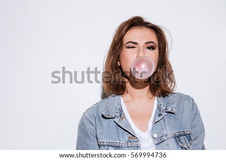 Image of a young playful lady dressed in jeans jacket standing isolated over white background while blowing bubble with chewing gum. Look at camera.