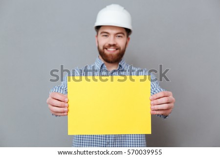 Photo of happy bearded man builder showing copyspace blank to camera standing over grey background. Focus on blank.