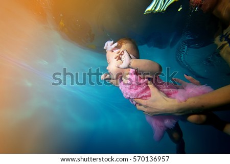 Happy baby in a beautiful dress swims underwater in the pool the rays of light, and his mother helps him. Portrait. Close-up. Shooting under water. Landscape orientation