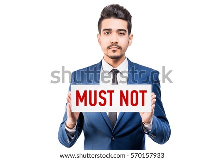 Young attractive man in a blue suit holding a white sign with red inscription MUST NOT on a white background. Isolated