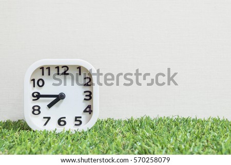 Closeup white clock for decorate show a quarter to eight or 7:45 a.m. on green artificial grass floor and cream wallpaper textured background with copy space