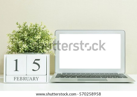 White wooden calendar with black 15 february word with white blank space at center of computer notebook on white wood desk and cream wallpaper textured background in selective focus at the calendar