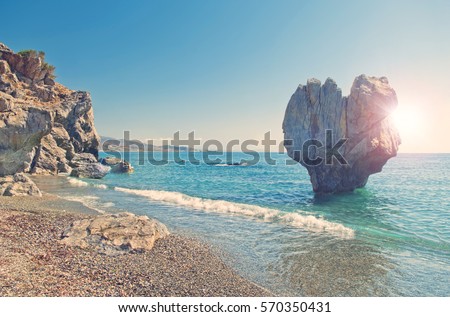 cross-processed image of huge heart shaped rock on pebble beach of Preveli with sun shining behind the rock, Crete, Greece