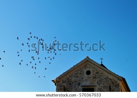 pigeons flying in the blue sky above a house build from stone with a crucifix on the roof/flock of flying doves,- freedom loving/birds, symbol for liberty and independence