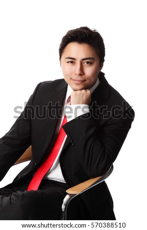 Sharp and handsome businessman in a black suit with red tie, sitting down on a chair in front of a white background, looking at camera.