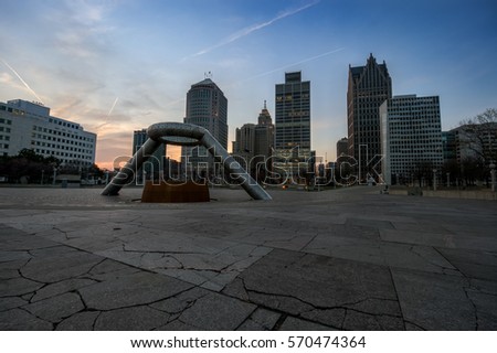 A square with a monument in a large metropolis with multiple tall buildings beneath blue skies 